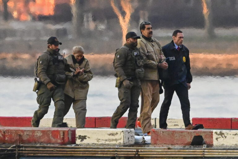 « Sur un quai en bord d’eau, quatre agents en tenue tactique — dont certains portent des blousons marqués “DEA” — escortent Nicolàs Maduro et Cilia Flores menottées vêtues de blousons et pantalons beiges. Le soleil couchant teinte le ciel d’orangé, et des blocs de béton peints en rouge et blanc délimitent le quai devant une surface d’eau calme. »