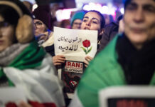 En Iran, le régime tue des milliers de manifestant-es Une manifestation nocturne : au centre, une jeune femme tient un panneau où figure un dessin de tulipe et un texte en persan, accompagné en dessous par l’inscription en anglais “IRAN : Total Communications BLACKOUT” et une carte stylisée de l’Iran barrée d’un symbole Wi-Fi. Autour d’elle, on devine d’autres manifestants et des drapeaux verts, blancs et rouges de l’Iran, dans une atmosphère urbaine éclairée.