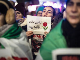 En Iran, le régime tue des milliers de manifestant-es Une manifestation nocturne : au centre, une jeune femme tient un panneau où figure un dessin de tulipe et un texte en persan, accompagné en dessous par l’inscription en anglais “IRAN : Total Communications BLACKOUT” et une carte stylisée de l’Iran barrée d’un symbole Wi-Fi. Autour d’elle, on devine d’autres manifestants et des drapeaux verts, blancs et rouges de l’Iran, dans une atmosphère urbaine éclairée.