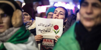En Iran, le régime tue des milliers de manifestant-es Une manifestation nocturne : au centre, une jeune femme tient un panneau où figure un dessin de tulipe et un texte en persan, accompagné en dessous par l’inscription en anglais “IRAN : Total Communications BLACKOUT” et une carte stylisée de l’Iran barrée d’un symbole Wi-Fi. Autour d’elle, on devine d’autres manifestants et des drapeaux verts, blancs et rouges de l’Iran, dans une atmosphère urbaine éclairée.