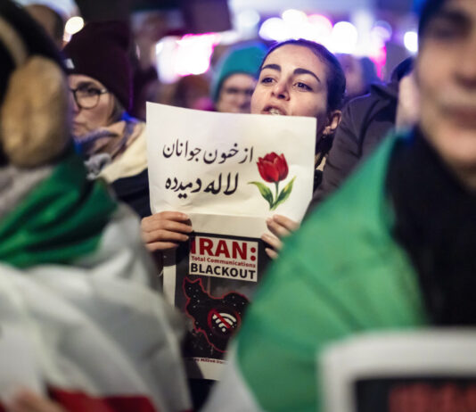 En Iran, le régime tue des milliers de manifestant-es Une manifestation nocturne : au centre, une jeune femme tient un panneau où figure un dessin de tulipe et un texte en persan, accompagné en dessous par l’inscription en anglais “IRAN : Total Communications BLACKOUT” et une carte stylisée de l’Iran barrée d’un symbole Wi-Fi. Autour d’elle, on devine d’autres manifestants et des drapeaux verts, blancs et rouges de l’Iran, dans une atmosphère urbaine éclairée.