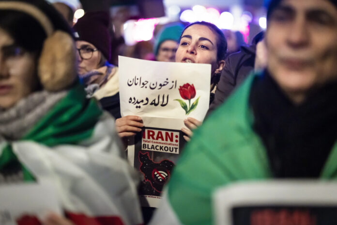 Une manifestation nocturne : au centre, une jeune femme tient un panneau où figure un dessin de tulipe et un texte en persan, accompagné en dessous par l’inscription en anglais “IRAN : Total Communications BLACKOUT” et une carte stylisée de l’Iran barrée d’un symbole Wi-Fi. Autour d’elle, on devine d’autres manifestants et des drapeaux verts, blancs et rouges de l’Iran, dans une atmosphère urbaine éclairée.