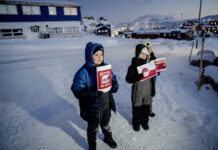 Le Groenland — pris entre le colonialisme et la politique des grandes puissances Trois enfants emmitouflés dans d’épaisses vestes et combinaisons d’hiver se tiennent sur une route enneigée au crépuscule, chacun brandissant une pancarte rouge-blanche. Au premier plan, un garçon tient une affiche « Greenland Not For Sale » avec un ours polaire stylisé. À ses côtés, deux autres enfants présentent des visuels inspirés du drapeau du Groenland. À l’arrière-plan, on devine quelques maisons aux fenêtres éclairées et les lumières d’un petit village sous la neige.