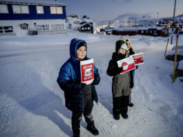 Le Groenland — pris entre le colonialisme et la politique des grandes puissances Trois enfants emmitouflés dans d’épaisses vestes et combinaisons d’hiver se tiennent sur une route enneigée au crépuscule, chacun brandissant une pancarte rouge-blanche. Au premier plan, un garçon tient une affiche « Greenland Not For Sale » avec un ours polaire stylisé. À ses côtés, deux autres enfants présentent des visuels inspirés du drapeau du Groenland. À l’arrière-plan, on devine quelques maisons aux fenêtres éclairées et les lumières d’un petit village sous la neige.