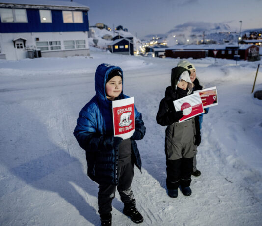 Le Groenland — pris entre le colonialisme et la politique des grandes puissances Trois enfants emmitouflés dans d’épaisses vestes et combinaisons d’hiver se tiennent sur une route enneigée au crépuscule, chacun brandissant une pancarte rouge-blanche. Au premier plan, un garçon tient une affiche « Greenland Not For Sale » avec un ours polaire stylisé. À ses côtés, deux autres enfants présentent des visuels inspirés du drapeau du Groenland. À l’arrière-plan, on devine quelques maisons aux fenêtres éclairées et les lumières d’un petit village sous la neige.