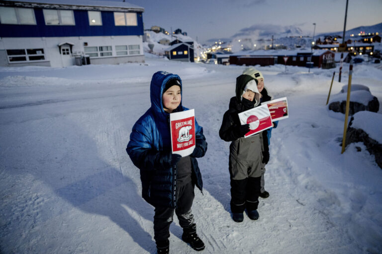 Trois enfants emmitouflés dans d’épaisses vestes et combinaisons d’hiver se tiennent sur une route enneigée au crépuscule, chacun brandissant une pancarte rouge-blanche. Au premier plan, un garçon tient une affiche « Greenland Not For Sale » avec un ours polaire stylisé. À ses côtés, deux autres enfants présentent des visuels inspirés du drapeau du Groenland. À l’arrière-plan, on devine quelques maisons aux fenêtres éclairées et les lumières d’un petit village sous la neige.