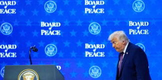 Le « Conseil de paix » : un rejet du droit international Donald Trump, un homme en costume sombre, avec une cravate, se tient devant un pupitre officiel. Le pupitre porte le sceau présidentiel des États-Unis. L’homme semble s’exprimer ou écouter attentivement, avec un micro devant lui. Derrière lui, un fond bleu présente à plusieurs reprises le texte « BOARD PEACE » ainsi que le logo du département d’État américain. Une petite composition florale blanche est placée sur le pupitre à droite.