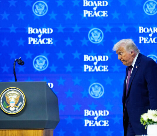 Le « Conseil de paix » : un rejet du droit international Donald Trump, un homme en costume sombre, avec une cravate, se tient devant un pupitre officiel. Le pupitre porte le sceau présidentiel des États-Unis. L’homme semble s’exprimer ou écouter attentivement, avec un micro devant lui. Derrière lui, un fond bleu présente à plusieurs reprises le texte « BOARD PEACE » ainsi que le logo du département d’État américain. Une petite composition florale blanche est placée sur le pupitre à droite.