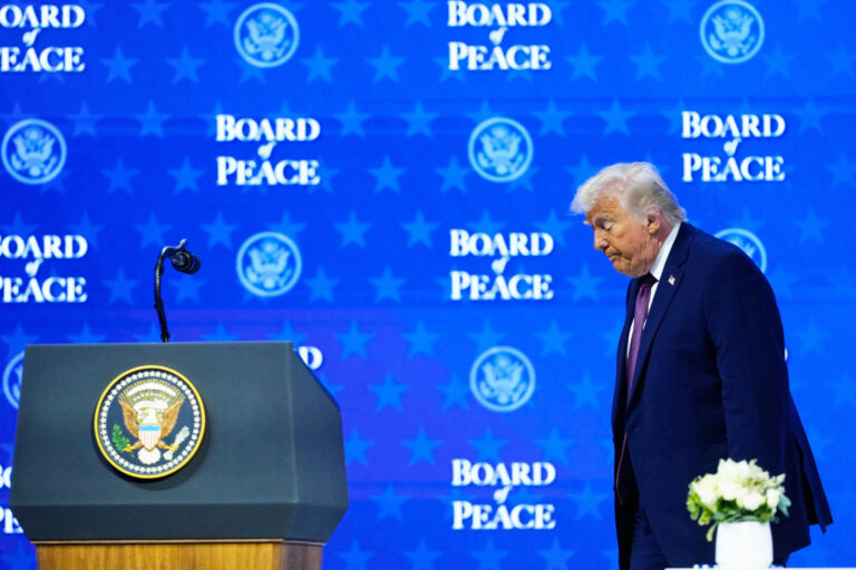 Donald Trump, un homme en costume sombre, avec une cravate, se tient devant un pupitre officiel. Le pupitre porte le sceau présidentiel des États-Unis. L’homme semble s’exprimer ou écouter attentivement, avec un micro devant lui. Derrière lui, un fond bleu présente à plusieurs reprises le texte « BOARD PEACE » ainsi que le logo du département d’État américain. Une petite composition florale blanche est placée sur le pupitre à droite.