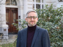 Manuel Puppis : « L’initiative entraînerait une réduction de moitié du budget annuel de la SSR » Manuel Puppis, un homme d’âge moyen, aux cheveux et à la barbe grisonnants, porte un col roulé sombre sous une veste de costume bleu foncé. Il sourit légèrement en regardant l’appareil. En arrière-plan, on aperçoit la façade blanche d’un bâtiment à l’architecture classique avec une porte en bois sculptée, ainsi qu’un taillis de buissons verts parsemés de petites baies rouges.