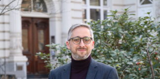 Manuel Puppis : « L’initiative entraînerait une réduction de moitié du budget annuel de la SSR » Manuel Puppis, un homme d’âge moyen, aux cheveux et à la barbe grisonnants, porte un col roulé sombre sous une veste de costume bleu foncé. Il sourit légèrement en regardant l’appareil. En arrière-plan, on aperçoit la façade blanche d’un bâtiment à l’architecture classique avec une porte en bois sculptée, ainsi qu’un taillis de buissons verts parsemés de petites baies rouges.