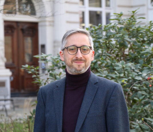 Manuel Puppis : « L’initiative entraînerait une réduction de moitié du budget annuel de la SSR » Manuel Puppis, un homme d’âge moyen, aux cheveux et à la barbe grisonnants, porte un col roulé sombre sous une veste de costume bleu foncé. Il sourit légèrement en regardant l’appareil. En arrière-plan, on aperçoit la façade blanche d’un bâtiment à l’architecture classique avec une porte en bois sculptée, ainsi qu’un taillis de buissons verts parsemés de petites baies rouges.