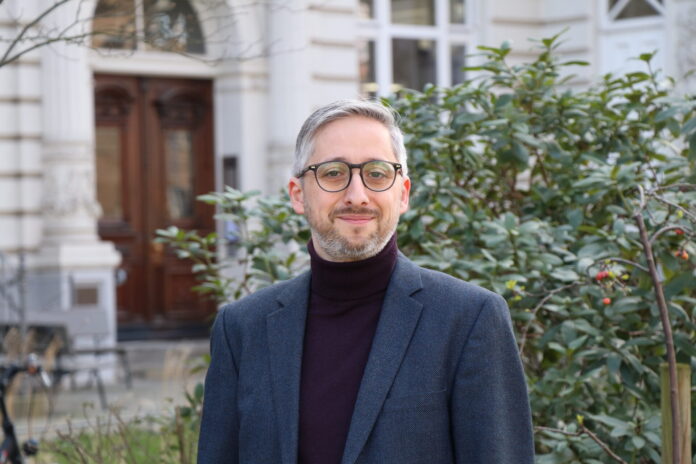Manuel Puppis, un homme d’âge moyen, aux cheveux et à la barbe grisonnants, porte un col roulé sombre sous une veste de costume bleu foncé. Il sourit légèrement en regardant l’appareil. En arrière-plan, on aperçoit la façade blanche d’un bâtiment à l’architecture classique avec une porte en bois sculptée, ainsi qu’un taillis de buissons verts parsemés de petites baies rouges.