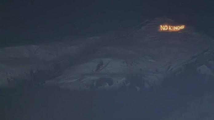Vue nocturne d’un versant montagneux sombre sous un ciel étoilé. Sur une crête à droite, un message lumineux en lettres incandescentes affiche « NO KINGS », se détachant nettement sur le relief obscur.