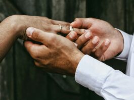 Imposition individuelle : le système fiscal qui favorise l’égalité Nahaufnahme zweier Hände: Eine Person in weissem Hemd steckt einer anderen, die bereits einen feinen Diamantring am Ringfinger trägt, einen schlichten silbernen Ring auf. Im unscharfen Hintergrund sind dunkelgraue Holzbretter zu erkennen.