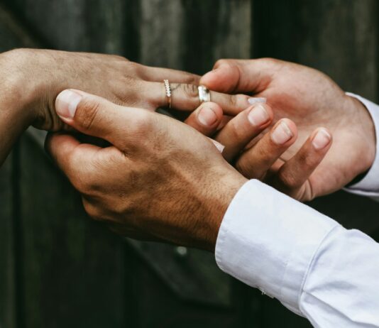 Imposition individuelle : le système fiscal qui favorise l’égalité Nahaufnahme zweier Hände: Eine Person in weissem Hemd steckt einer anderen, die bereits einen feinen Diamantring am Ringfinger trägt, einen schlichten silbernen Ring auf. Im unscharfen Hintergrund sind dunkelgraue Holzbretter zu erkennen.