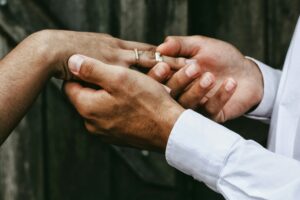 Gros plan sur deux mains : une main vêtue d’une chemise blanche tient délicatement le doigt d’une autre personne — déjà ornée d’une fine bague sertie de petits diamants — et y glisse un anneau d’or massif. L’arrière-plan est flou, créant une atmosphère intime et concentrant l’attention sur l’échange de bagues.