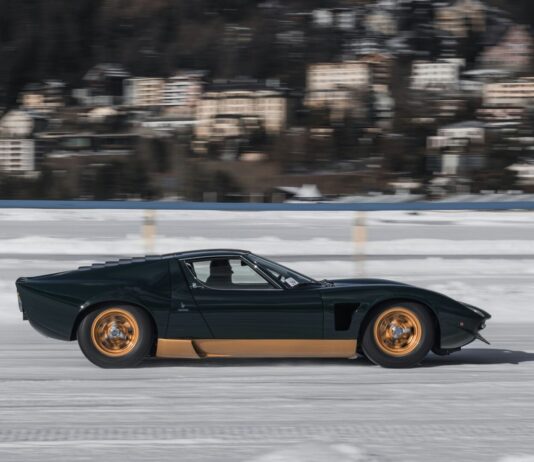 La richesse disproportionnée menace la démocratie vue de profil d’une voiture de sport classique à moteur central, de teinte vert foncé, jantes dorées, filant à vive allure sur une surface gelée et enneigée. À l’arrière-plan, un village aux façades pastel apparaît flou, soulignant l’effet de vitesse.