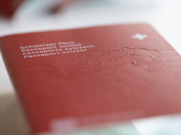 La naturalisation en Suisse : arbitraire et discriminatoire Gros plan sur la couverture rouge d’un passeport suisse posé à plat. On distingue au premier plan, en lettres blanches et légèrement floues, les inscriptions en allemand, français, italien et romanche (« Schweizer Pass / Passeport suisse / Passaporto svizzero / Passaport svizzer ») ainsi qu’une petite croix blanche en haut à droite. Le relief gravé du massif alpin est perceptible sous la surface texturée du document. Le fond et le bord inférieur sont hors de focus, soulignant la profondeur de champ réduite.