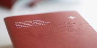 La naturalisation en Suisse : arbitraire et discriminatoire Gros plan sur la couverture rouge d’un passeport suisse posé à plat. On distingue au premier plan, en lettres blanches et légèrement floues, les inscriptions en allemand, français, italien et romanche (« Schweizer Pass / Passeport suisse / Passaporto svizzero / Passaport svizzer ») ainsi qu’une petite croix blanche en haut à droite. Le relief gravé du massif alpin est perceptible sous la surface texturée du document. Le fond et le bord inférieur sont hors de focus, soulignant la profondeur de champ réduite.