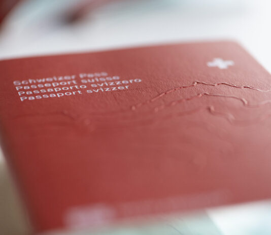 La naturalisation en Suisse : arbitraire et discriminatoire Gros plan sur la couverture rouge d’un passeport suisse posé à plat. On distingue au premier plan, en lettres blanches et légèrement floues, les inscriptions en allemand, français, italien et romanche (« Schweizer Pass / Passeport suisse / Passaporto svizzero / Passaport svizzer ») ainsi qu’une petite croix blanche en haut à droite. Le relief gravé du massif alpin est perceptible sous la surface texturée du document. Le fond et le bord inférieur sont hors de focus, soulignant la profondeur de champ réduite.