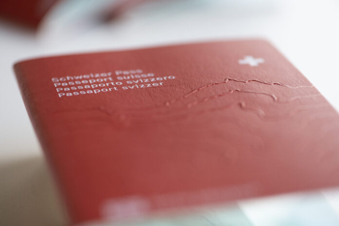 Gros plan sur la couverture rouge d’un passeport suisse posé à plat. On distingue au premier plan, en lettres blanches et légèrement floues, les inscriptions en allemand, français, italien et romanche (« Schweizer Pass / Passeport suisse / Passaporto svizzero / Passaport svizzer ») ainsi qu’une petite croix blanche en haut à droite. Le relief gravé du massif alpin est perceptible sous la surface texturée du document. Le fond et le bord inférieur sont hors de focus, soulignant la profondeur de champ réduite.