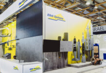 Une entreprise suisse fournit une technologie laser à un groupe d’armement israélien Vue d’un stand d’exposition de la société Elbit Systems dans un vaste hall aux structures en bois apparentes. Le fond du stand est peint en blanc et jaune, orné du logo “Elbit Systems”. À gauche sont suspendues des maquettes de drones et d’aéronefs, au centre un grand écran noir trône au-dessus de deux bombes suspendues, et à droite plusieurs modèles grandeur nature de bombes et de missiles sont alignés sur un socle. Devant, un comptoir d’accueil blanc arbore également le logo de la marque, tandis que deux visiteurs sont assis sur la droite.