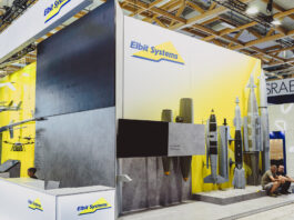 Une entreprise suisse fournit une technologie laser à un groupe d’armement israélien Vue d’un stand d’exposition de la société Elbit Systems dans un vaste hall aux structures en bois apparentes. Le fond du stand est peint en blanc et jaune, orné du logo “Elbit Systems”. À gauche sont suspendues des maquettes de drones et d’aéronefs, au centre un grand écran noir trône au-dessus de deux bombes suspendues, et à droite plusieurs modèles grandeur nature de bombes et de missiles sont alignés sur un socle. Devant, un comptoir d’accueil blanc arbore également le logo de la marque, tandis que deux visiteurs sont assis sur la droite.
