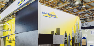 Une entreprise suisse fournit une technologie laser à un groupe d’armement israélien Vue d’un stand d’exposition de la société Elbit Systems dans un vaste hall aux structures en bois apparentes. Le fond du stand est peint en blanc et jaune, orné du logo “Elbit Systems”. À gauche sont suspendues des maquettes de drones et d’aéronefs, au centre un grand écran noir trône au-dessus de deux bombes suspendues, et à droite plusieurs modèles grandeur nature de bombes et de missiles sont alignés sur un socle. Devant, un comptoir d’accueil blanc arbore également le logo de la marque, tandis que deux visiteurs sont assis sur la droite.