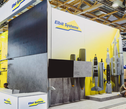Une entreprise suisse fournit une technologie laser à un groupe d’armement israélien Vue d’un stand d’exposition de la société Elbit Systems dans un vaste hall aux structures en bois apparentes. Le fond du stand est peint en blanc et jaune, orné du logo “Elbit Systems”. À gauche sont suspendues des maquettes de drones et d’aéronefs, au centre un grand écran noir trône au-dessus de deux bombes suspendues, et à droite plusieurs modèles grandeur nature de bombes et de missiles sont alignés sur un socle. Devant, un comptoir d’accueil blanc arbore également le logo de la marque, tandis que deux visiteurs sont assis sur la droite.