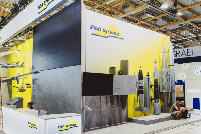 Vue d’un stand d’exposition de la société Elbit Systems dans un vaste hall aux structures en bois apparentes. Le fond du stand est peint en blanc et jaune, orné du logo “Elbit Systems”. À gauche sont suspendues des maquettes de drones et d’aéronefs, au centre un grand écran noir trône au-dessus de deux bombes suspendues, et à droite plusieurs modèles grandeur nature de bombes et de missiles sont alignés sur un socle. Devant, un comptoir d’accueil blanc arbore également le logo de la marque, tandis que deux visiteurs sont assis sur la droite.