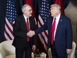 Trump et les droits de douane : la Suisse est dorénavant désavantagée Deux hommes, Guy Parmelin et Donald Trump, en costume-cravate se serrent la main devant plusieurs drapeaux (États-Unis et Suisse). L’un porte un costume bleu foncé et une cravate rouge unie, l’autre un costume sombre et une cravate rouge à petits motifs. À l’arrière-plan, deux fauteuils blancs et une petite table basse ornée d’un bouquet et de verres.