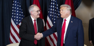 Trump et les droits de douane : la Suisse est dorénavant désavantagée Deux hommes, Guy Parmelin et Donald Trump, en costume-cravate se serrent la main devant plusieurs drapeaux (États-Unis et Suisse). L’un porte un costume bleu foncé et une cravate rouge unie, l’autre un costume sombre et une cravate rouge à petits motifs. À l’arrière-plan, deux fauteuils blancs et une petite table basse ornée d’un bouquet et de verres.