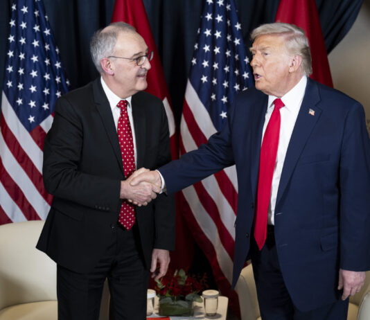 Trump et les droits de douane : la Suisse est dorénavant désavantagée Deux hommes, Guy Parmelin et Donald Trump, en costume-cravate se serrent la main devant plusieurs drapeaux (États-Unis et Suisse). L’un porte un costume bleu foncé et une cravate rouge unie, l’autre un costume sombre et une cravate rouge à petits motifs. À l’arrière-plan, deux fauteuils blancs et une petite table basse ornée d’un bouquet et de verres.