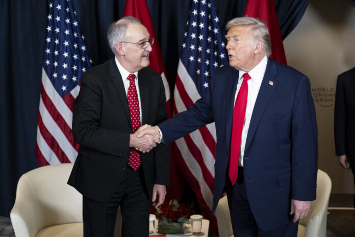 Deux hommes, Guy Parmelin et Donald Trump, en costume-cravate se serrent la main devant plusieurs drapeaux (États-Unis et Suisse). L’un porte un costume bleu foncé et une cravate rouge unie, l’autre un costume sombre et une cravate rouge à petits motifs. À l’arrière-plan, deux fauteuils blancs et une petite table basse ornée d’un bouquet et de verres.