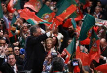 Bonnes nouvelles en février : élections au Portugal, interdiction de jeter les vêtements et reconnaissance des femmes transgenres Un homme en costume sombre, vu de profil, lève la main pour saluer une foule de partisans qui agitent de nombreux drapeaux portugais rouges et verts. Autour de lui, des visages enthousiastes, des micros et des appareils photo témoignent d’une ambiance de rassemblement politique.