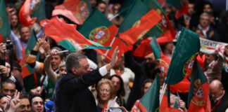 Bonnes nouvelles en février : élections au Portugal, interdiction de jeter les vêtements et reconnaissance des femmes transgenres Un homme en costume sombre, vu de profil, lève la main pour saluer une foule de partisans qui agitent de nombreux drapeaux portugais rouges et verts. Autour de lui, des visages enthousiastes, des micros et des appareils photo témoignent d’une ambiance de rassemblement politique.