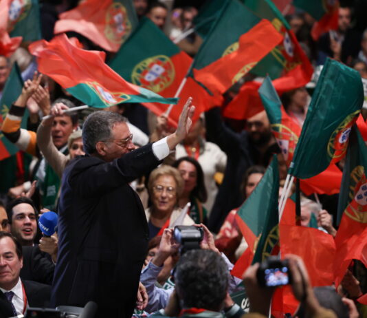 Bonnes nouvelles en février : élections au Portugal, interdiction de jeter les vêtements et reconnaissance des femmes transgenres Un homme en costume sombre, vu de profil, lève la main pour saluer une foule de partisans qui agitent de nombreux drapeaux portugais rouges et verts. Autour de lui, des visages enthousiastes, des micros et des appareils photo témoignent d’une ambiance de rassemblement politique.