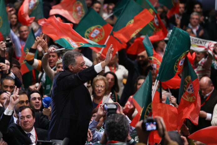 Un homme en costume sombre, vu de profil, lève la main pour saluer une foule de partisans qui agitent de nombreux drapeaux portugais rouges et verts. Autour de lui, des visages enthousiastes, des micros et des appareils photo témoignent d’une ambiance de rassemblement politique.