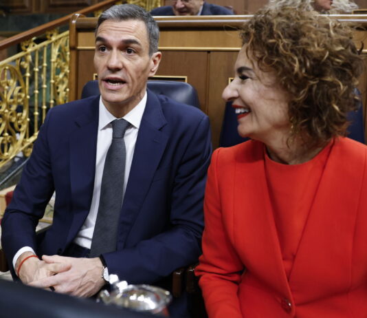 Contre-courant humain : l’Espagne veut régulariser un demi-million de migrant-es Espagne : deux personnes assises côte à côte dans un hémicycle aux boiseries et rambardes dorées. À gauche, Pedro Sánchez, Premier ministre espagnol, un homme en costume bleu foncé, chemise blanche et cravate grise, semble prendre la parole. À droite, sa première vice-première ministre, Maria Jesus Montero, une femme portant un tailleur rouge sourit en le regardant.