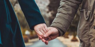 Pourquoi il est grand temps d’introduire l’imposition individuelle Deux personnes main dans la main au centre d’une route de campagne : à gauche, une silhouette vêtue d’un manteau vert foncé, à droite, une autre portant un parka kaki, avec en arrière-plan un paysage flou de collines et d’arbres.