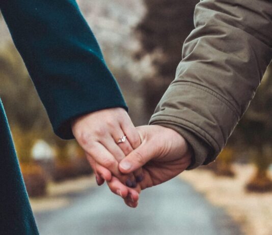 Pourquoi il est grand temps d’introduire l’imposition individuelle Deux personnes main dans la main au centre d’une route de campagne : à gauche, une silhouette vêtue d’un manteau vert foncé, à droite, une autre portant un parka kaki, avec en arrière-plan un paysage flou de collines et d’arbres.