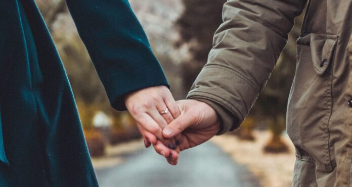 Deux personnes main dans la main au centre d’une route de campagne : à gauche, une silhouette vêtue d’un manteau vert foncé, à droite, une autre portant un parka kaki, avec en arrière-plan un paysage flou de collines et d’arbres.