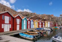 Semaine de 4 jours en Suède : des employé-es sont plus satisfait-es et une productivité plus élevée Vue d’un quai longeant des cabanes de pêcheurs colorées – rouges, bleu turquoise, jaune et blanc – alignées devant un rocher escarpé. Plusieurs petites embarcations à moteur et à rames sont amarrées dans l’eau calme, sous un ciel bleu parsemé de nuages.