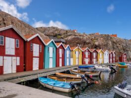 Semaine de 4 jours en Suède : des employé-es plus satisfait-es et une productivité plus élevée Vue d’un quai longeant des cabanes de pêcheurs colorées – rouges, bleu turquoise, jaune et blanc – alignées devant un rocher escarpé. Plusieurs petites embarcations à moteur et à rames sont amarrées dans l’eau calme, sous un ciel bleu parsemé de nuages.