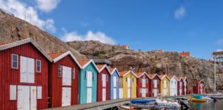 Semaine de 4 jours en Suède : des employé-es sont plus satisfait-es et une productivité plus élevée Vue d’un quai longeant des cabanes de pêcheurs colorées – rouges, bleu turquoise, jaune et blanc – alignées devant un rocher escarpé. Plusieurs petites embarcations à moteur et à rames sont amarrées dans l’eau calme, sous un ciel bleu parsemé de nuages.