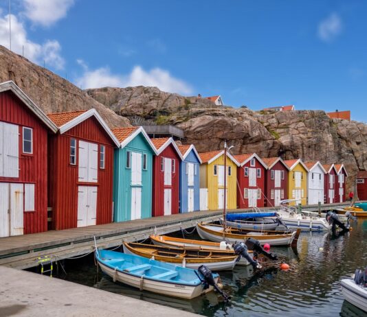 Semaine de 4 jours en Suède : des employé-es sont plus satisfait-es et une productivité plus élevée Vue d’un quai longeant des cabanes de pêcheurs colorées – rouges, bleu turquoise, jaune et blanc – alignées devant un rocher escarpé. Plusieurs petites embarcations à moteur et à rames sont amarrées dans l’eau calme, sous un ciel bleu parsemé de nuages.