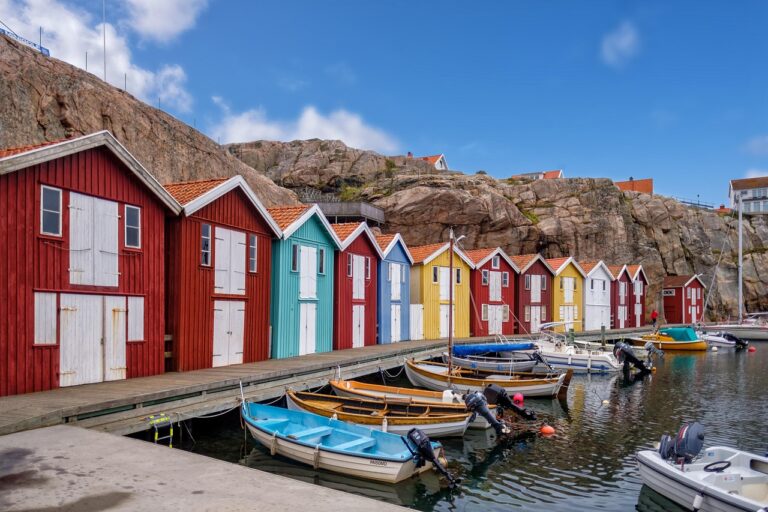 Vue d’un quai longeant des cabanes de pêcheurs colorées – rouges, bleu turquoise, jaune et blanc – alignées devant un rocher escarpé. Plusieurs petites embarcations à moteur et à rames sont amarrées dans l’eau calme, sous un ciel bleu parsemé de nuages.