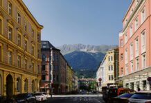 Autriche : un frein aux loyers pour tous-tes Rue dégagée en Autriche bordée de bâtiments anciens aux façades colorées – jaune, rose et ocre – avec des voitures stationnées le long du trottoir. Au fond, une chaîne de montagnes vertes coiffée de crêtes rocheuses se détache sous un ciel bleu clair. Sur la chaussée, le mot « STOP » est peint devant un carrefour signalé par un feu tricolore.