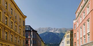 Autriche : un frein aux loyers pour tous-tes Rue dégagée en Autriche bordée de bâtiments anciens aux façades colorées – jaune, rose et ocre – avec des voitures stationnées le long du trottoir. Au fond, une chaîne de montagnes vertes coiffée de crêtes rocheuses se détache sous un ciel bleu clair. Sur la chaussée, le mot « STOP » est peint devant un carrefour signalé par un feu tricolore.